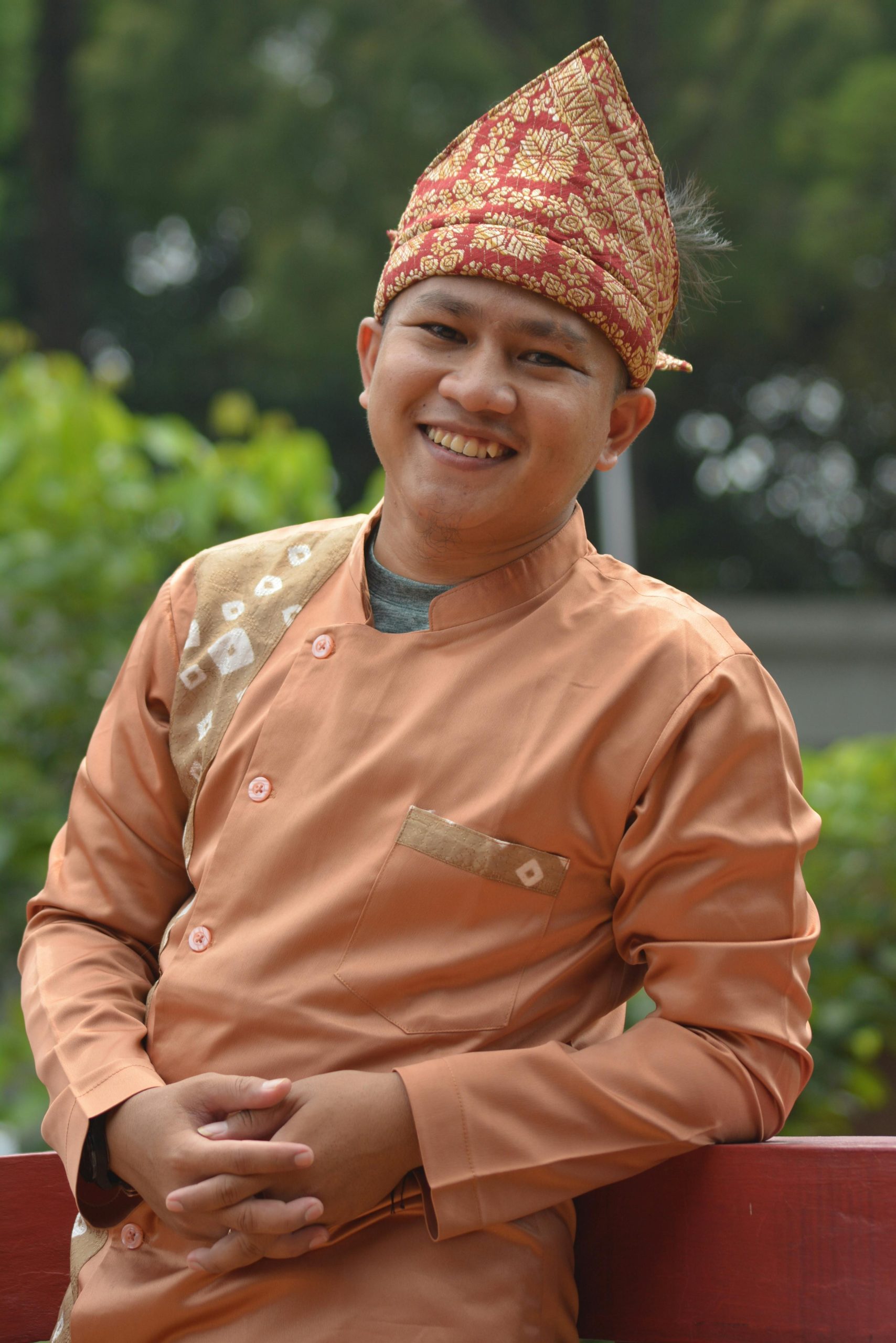 Cheerful man wearing traditional Indonesian clothing with turban, outside on a sunny day.